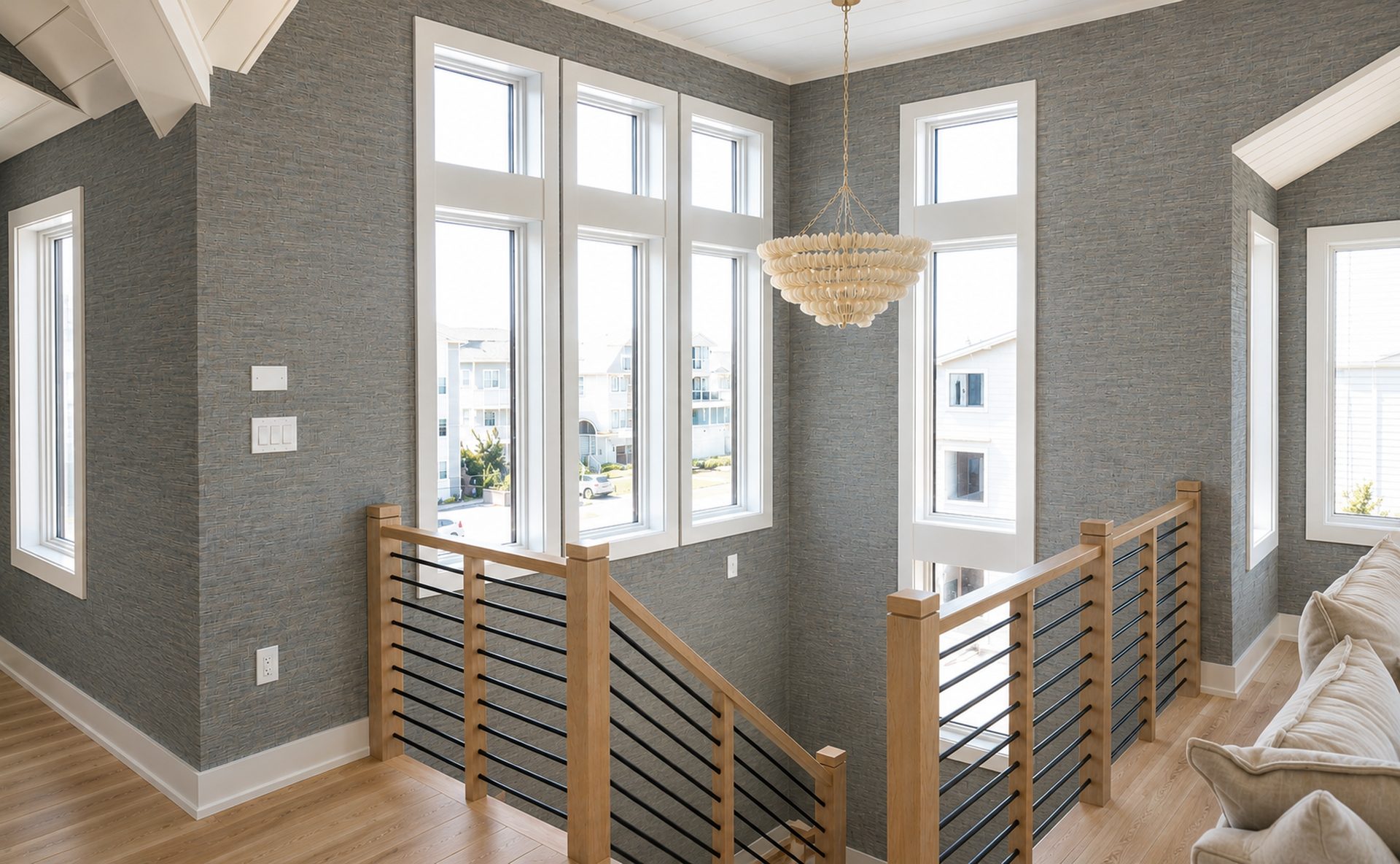 Textured grey grasscloth wallcovering installed by RGT Wallpaper across a sunlit two-storey stair landing with white oak railings and a beaded chandelier